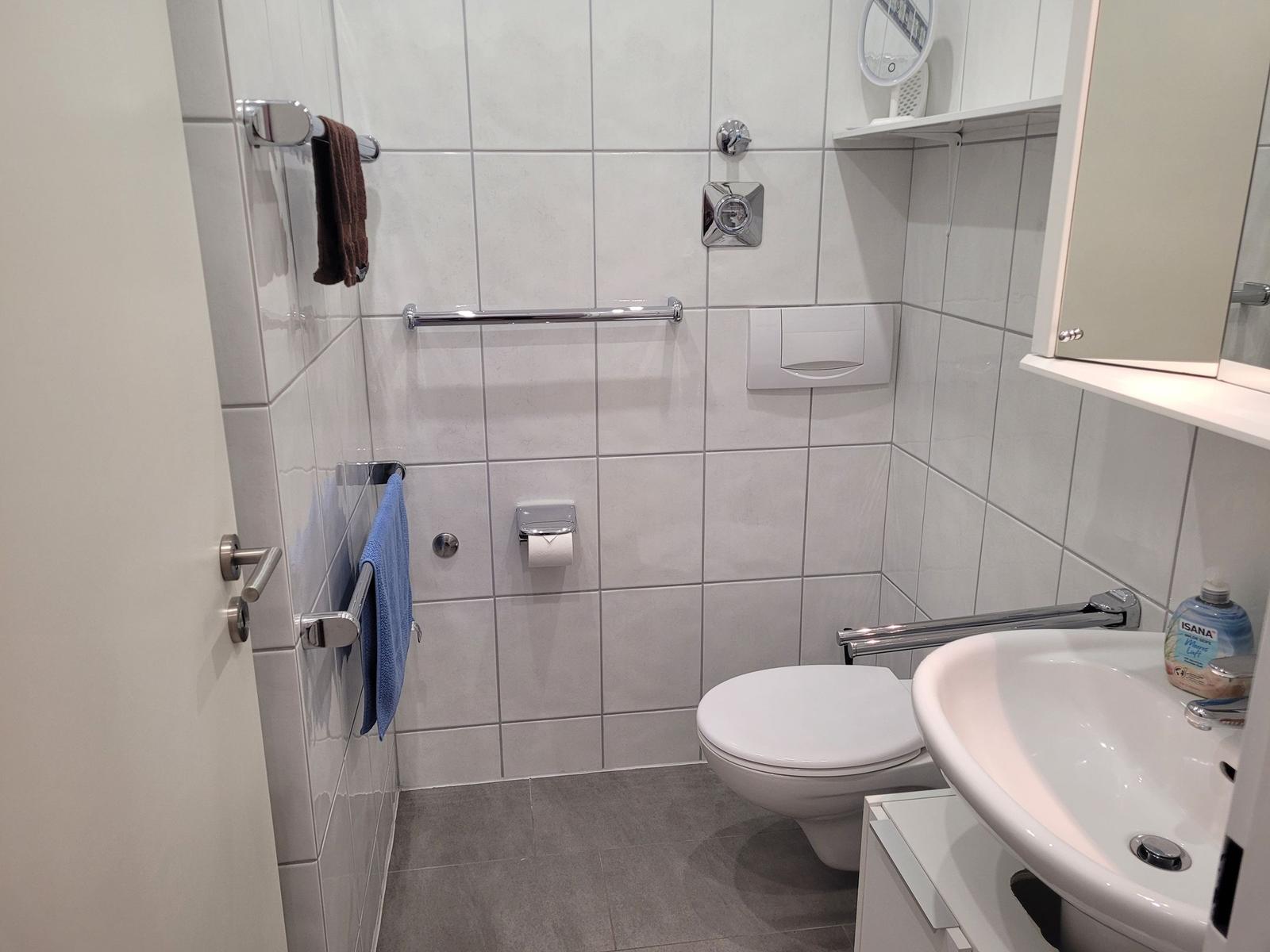 Badezimmer mit Toilette, Waschbecken und Handtuchhaltern. Fliesenwand und grauer Boden.