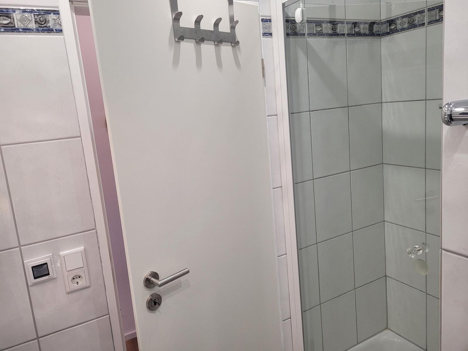 Dusche mit Glaswand, weiße Tür mit Haken und Fliesen mit Mosaikverzierung.