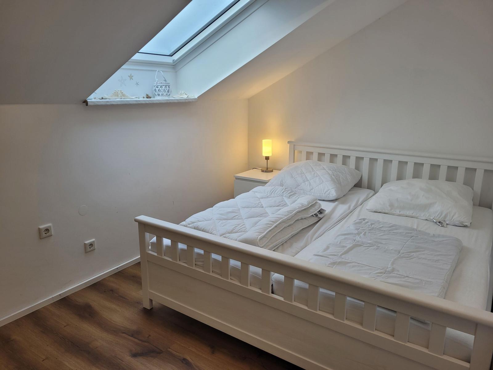Schlafzimmer mit Dachfenster, weißem Bett und Nachttisch mit Lampe.