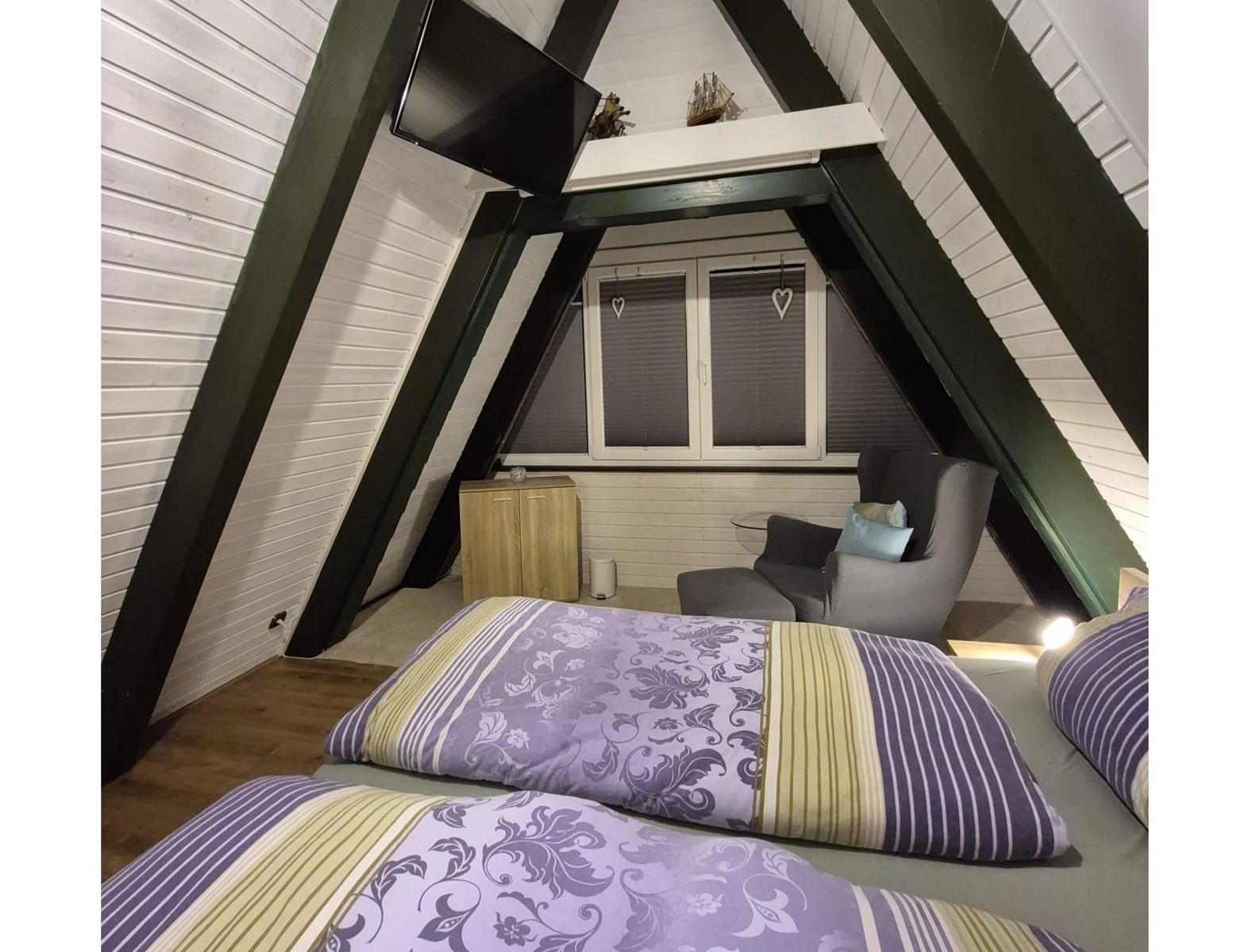 Dachgeschosszimmer mit Bett, Sessel und Fenster unter Holzdecke.