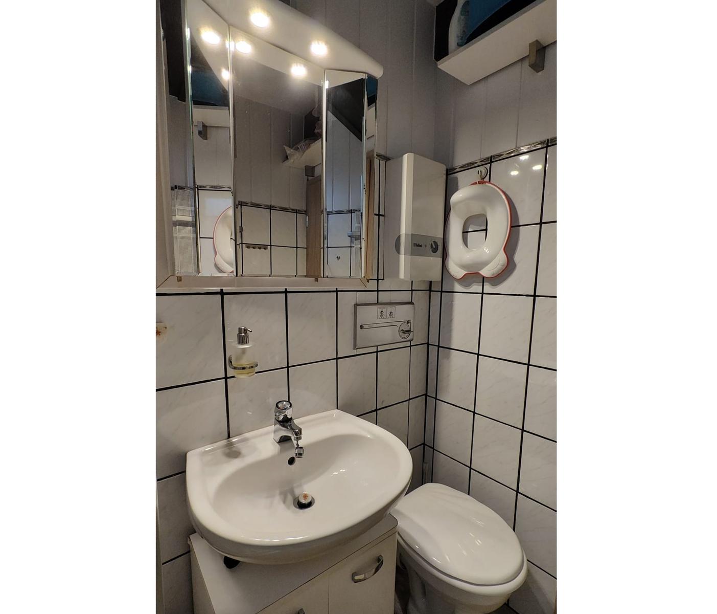 Badezimmer mit Waschbecken, Spiegelschrank und Toilette. Wandfliesen und Duschbereich sichtbar.