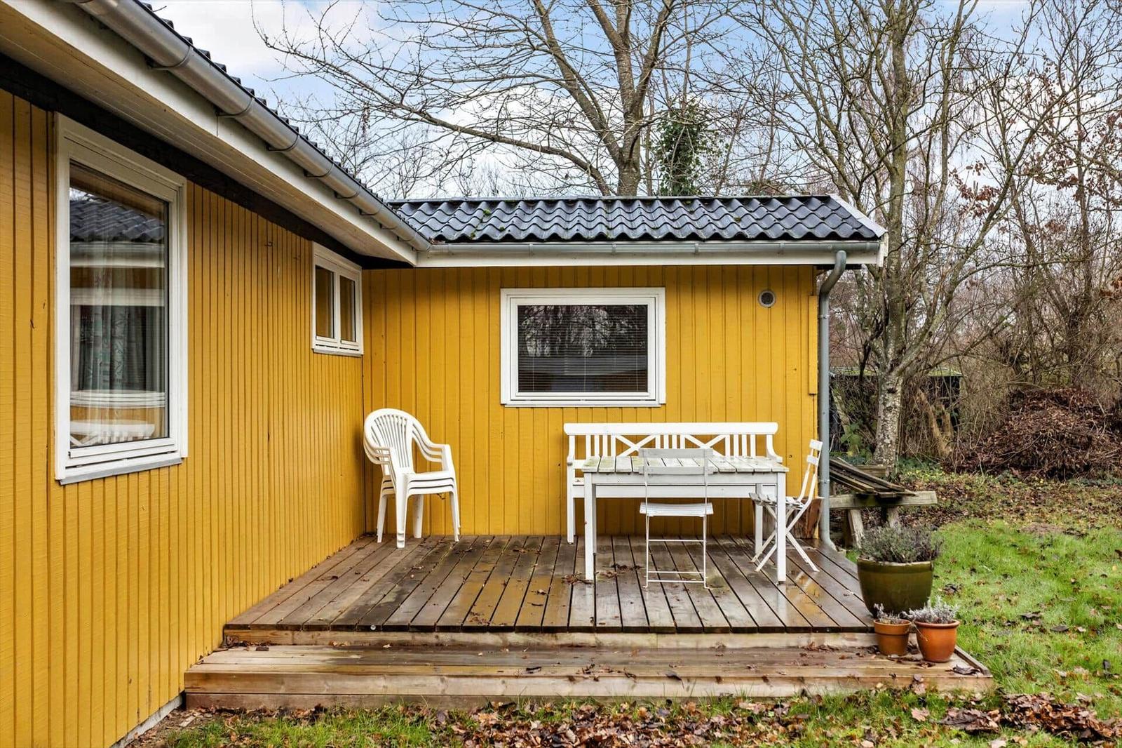 Yellow house with wooden terrace, white table and chairs, surrounded by trees.