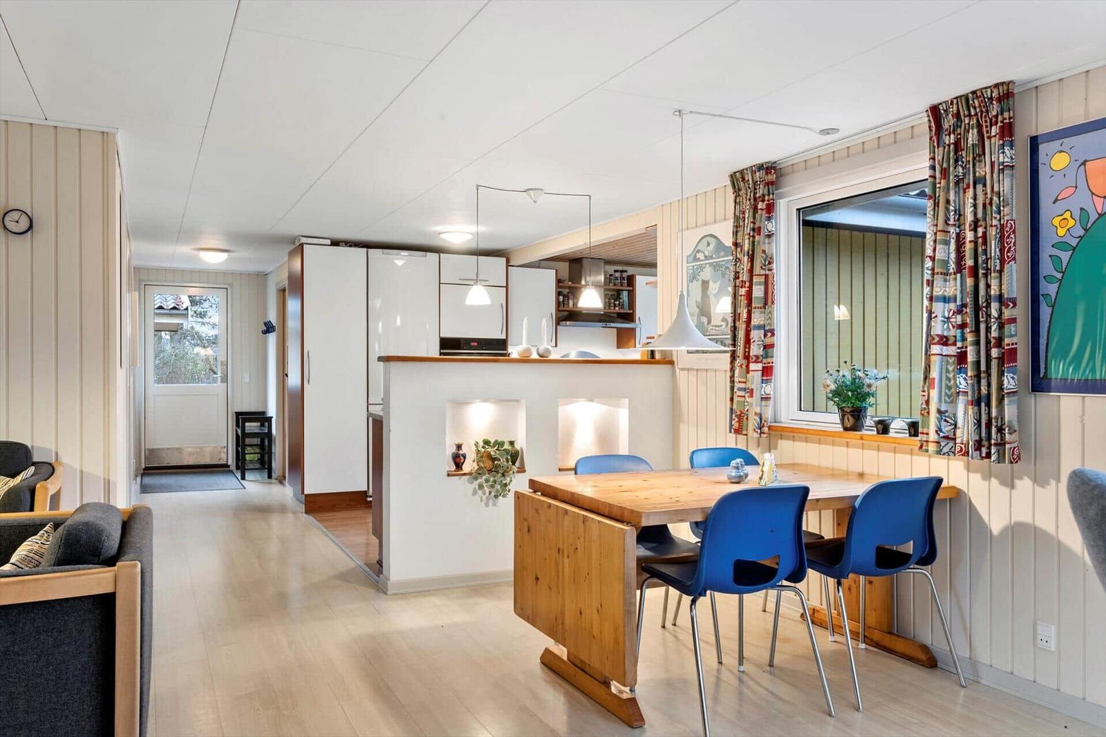 Dining area with wooden table and blue chairs, kitchen with white cabinets and window with colorful curtains.