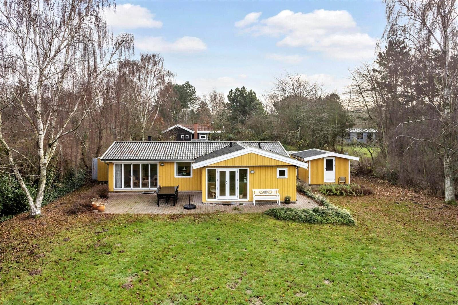 Yellow vacation house with terrace, garden, and trees. White bench and table are outside.