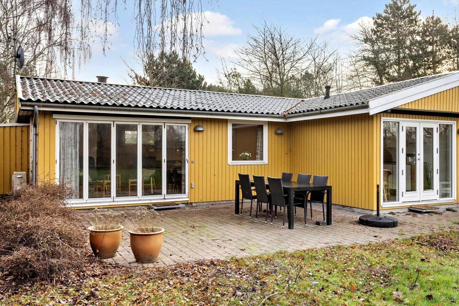 Yellow holiday home with terrace, table and chairs. Backyard with trees and plants.