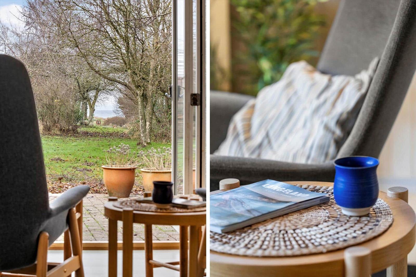 A gray chair with cushions and table with blue mug and book. View of garden and woods.