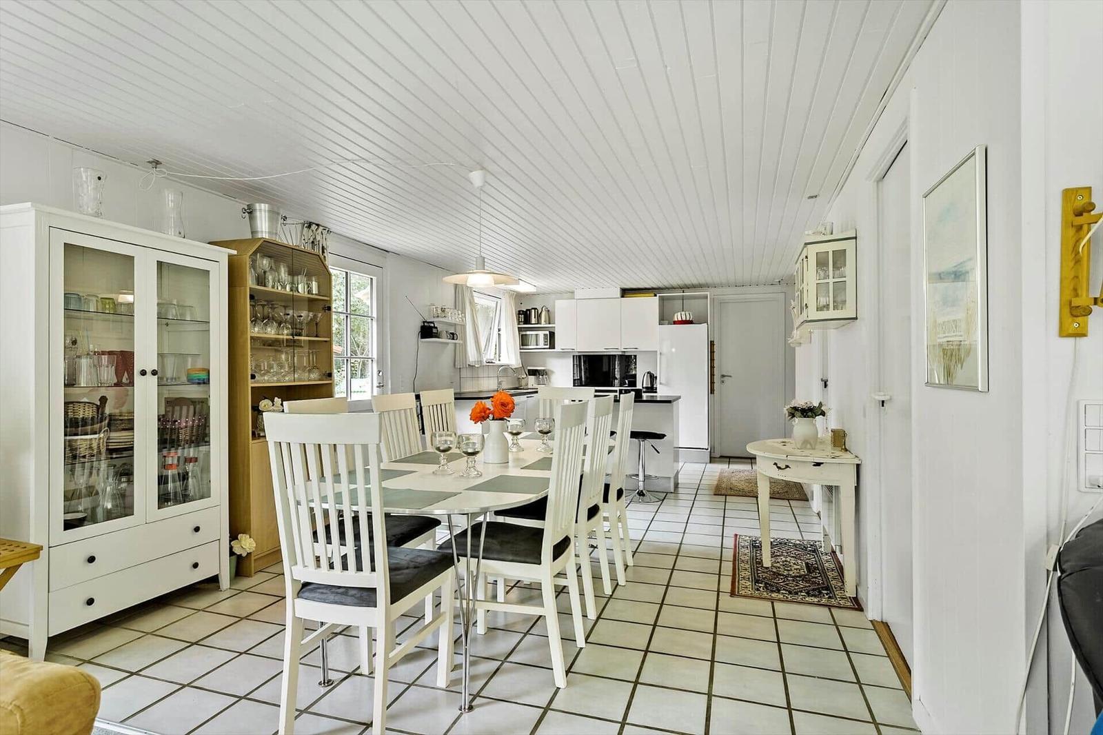 Dining area with table and chairs, kitchen with worktop and cabinets, white walls and floor tiles.