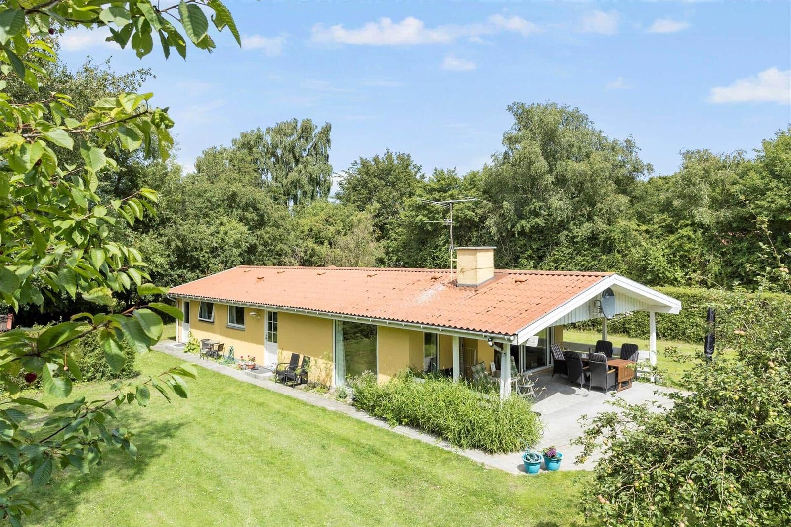 A yellow house with terrace and garden. Surrounded by trees under blue sky.
