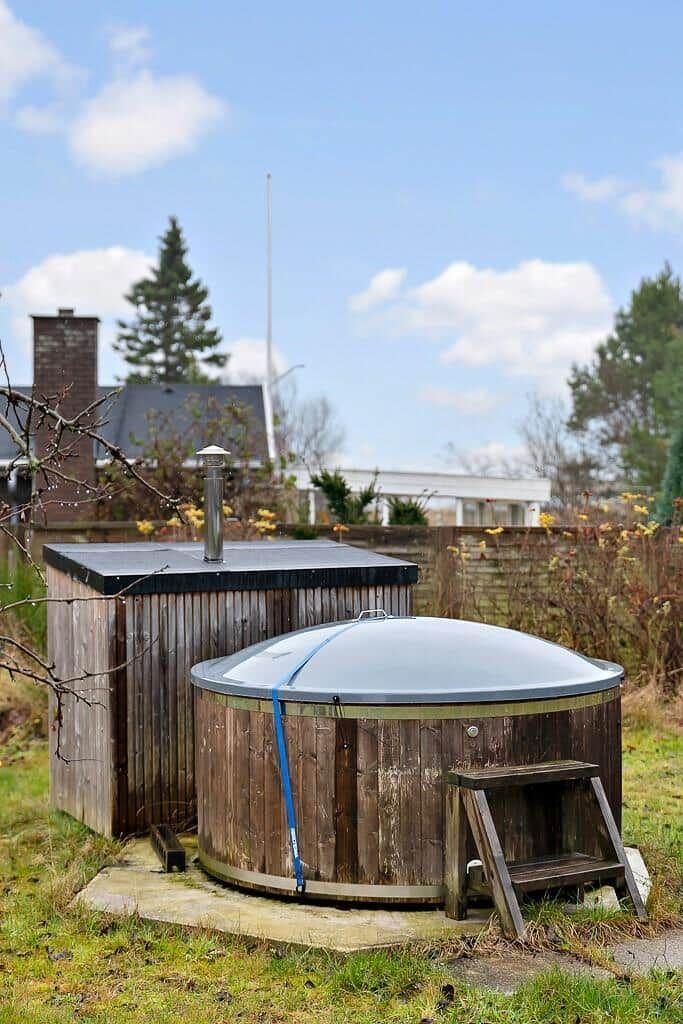 Wooden structure with roof and chimney, next to round wooden pool with metal lid and wooden step.