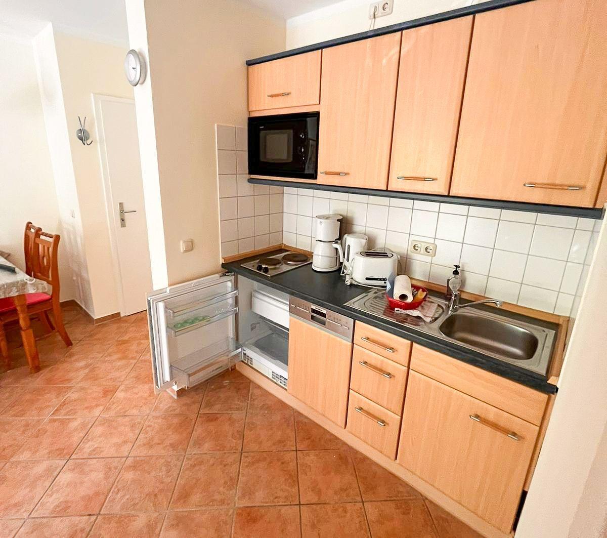 Küche mit Holzschränken, Spüle und Kühlschrank. Im Hintergrund ist Essbereich mit Tisch und Stühlen.
