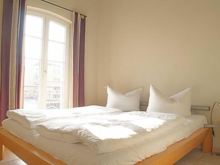 Ein Bett mit weißer Bettwäsche und zwei Kissen. Ein Fenster mit Vorhängen und Blick auf Balkon.