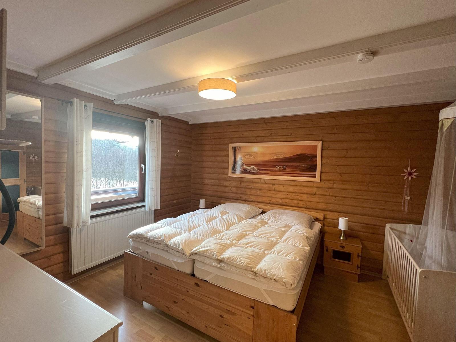 Bedroom with wooden wall, bed, crib, and window to the forest.