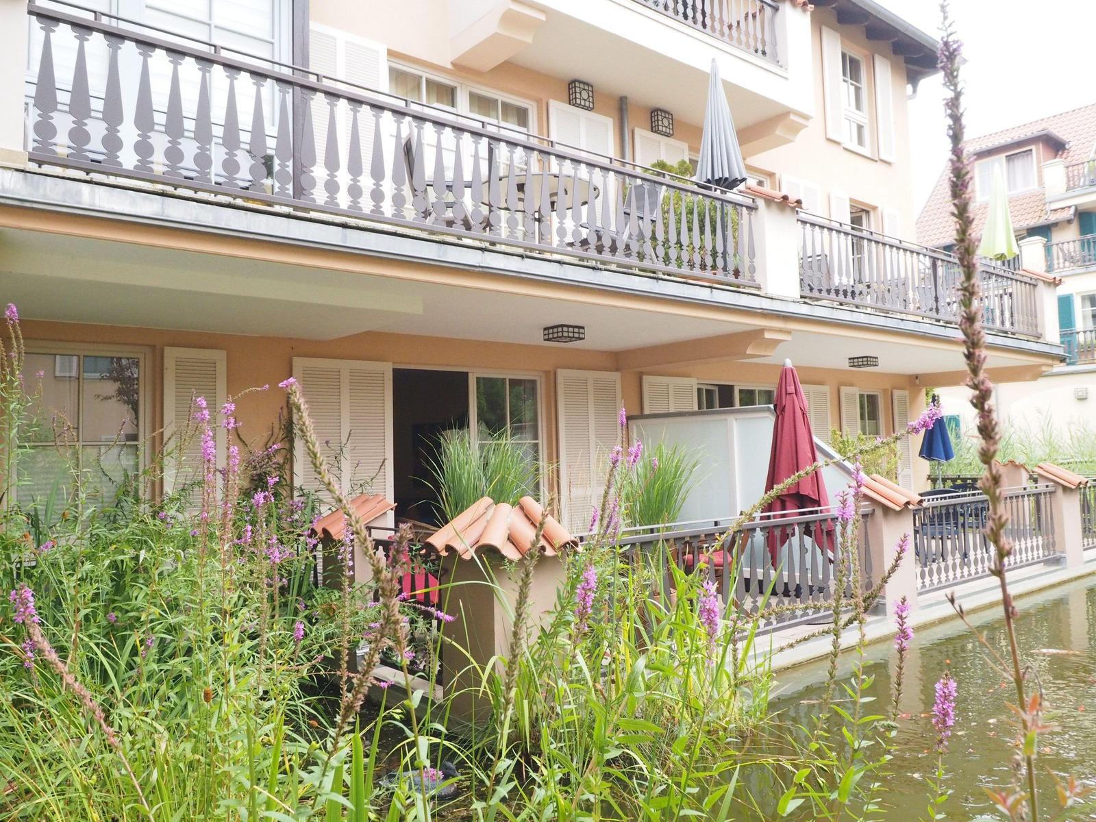 Haus mit Balkon und Garten am Wasser. Rote Sonnenschirme und Pflanzen sind sichtbar.