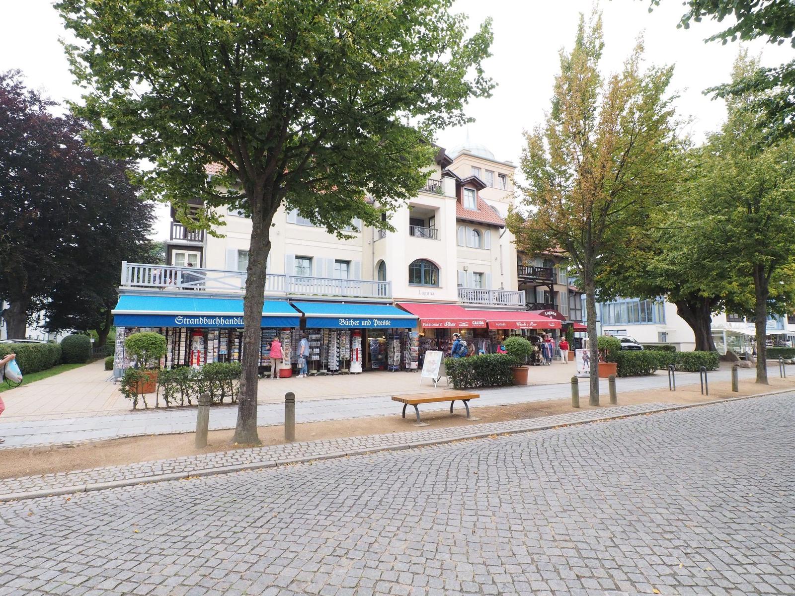 Strandbuchhandlung und Geschäfte am gepflasterten Weg mit Bäumen und einem Sitzbänkchen.