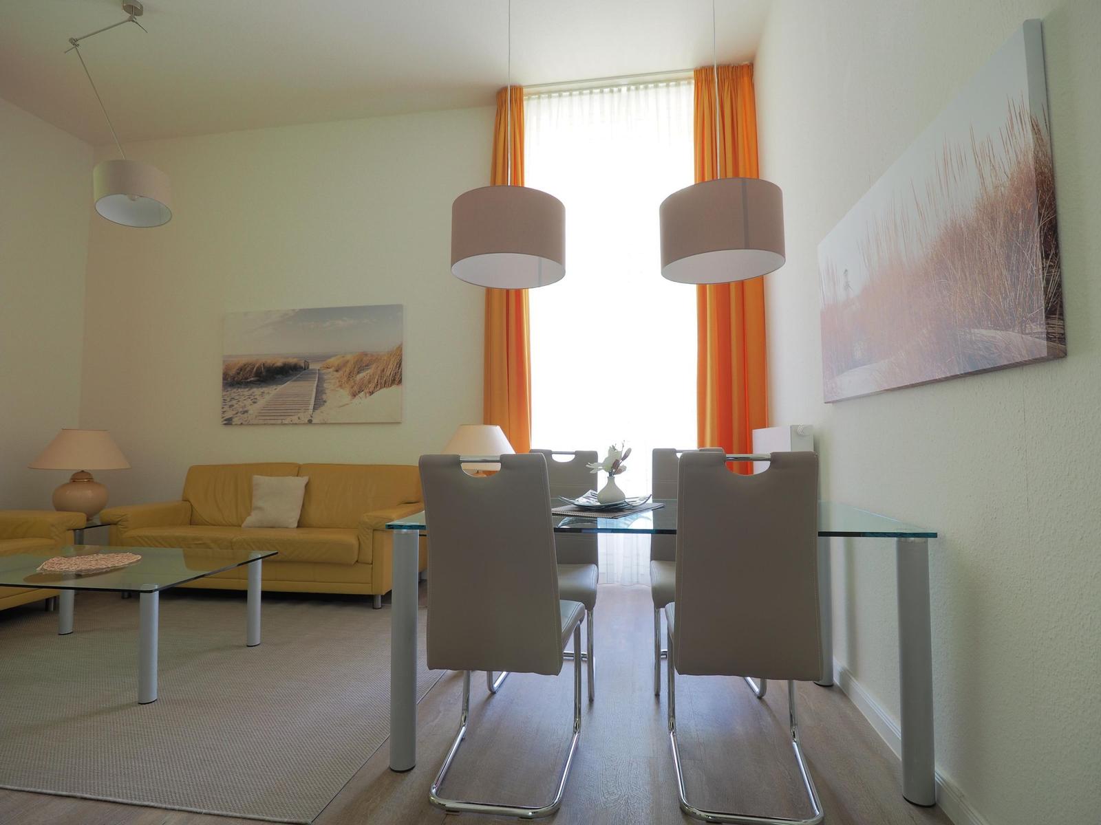 Wohn- und Essbereich mit Sofa, Tisch und Stühlen sowie Fenster mit orangefarbenen Vorhängen.