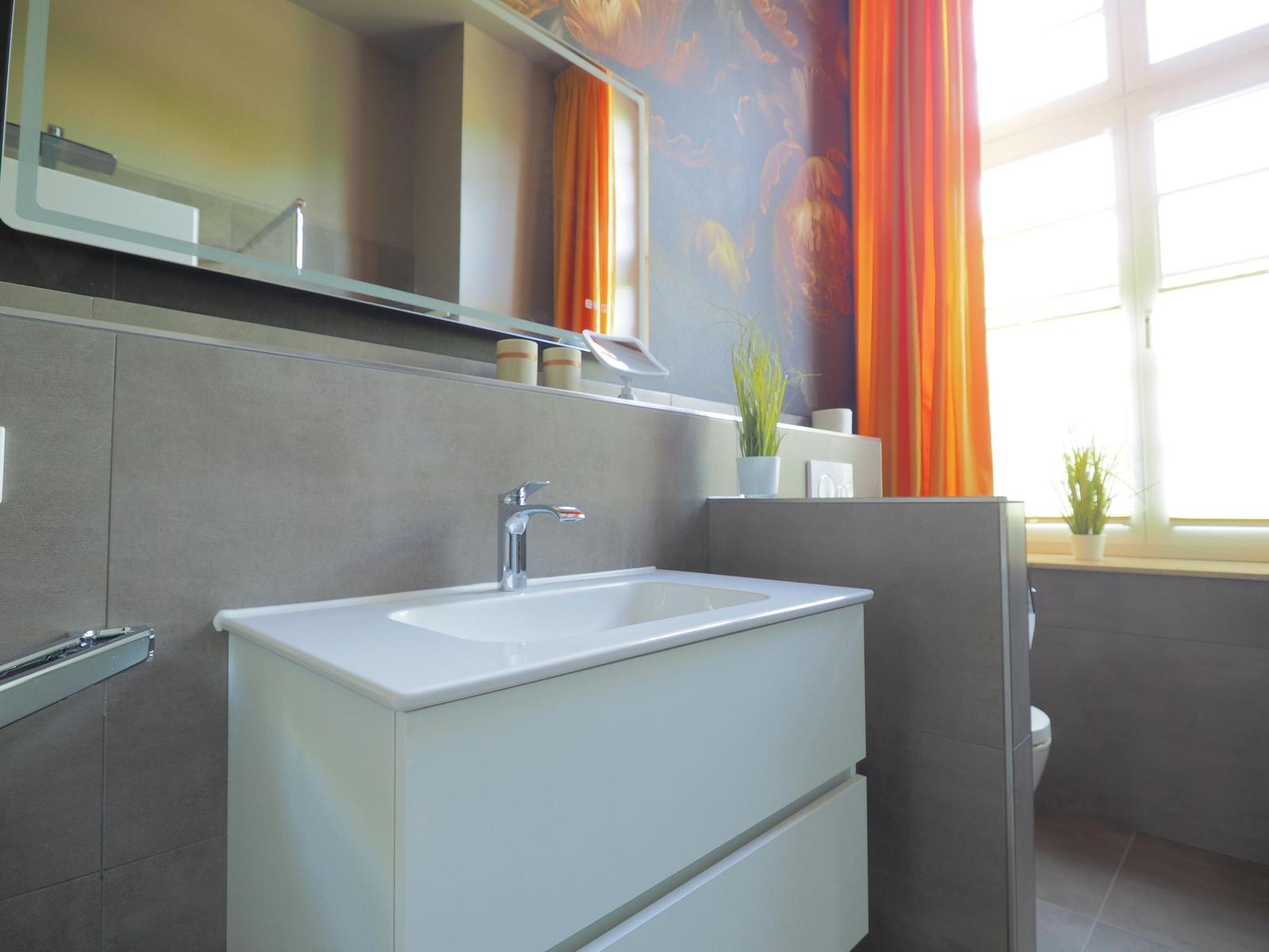 Bad mit Waschbecken, Spiegel und Fenster mit orangefarbenen Vorhängen.