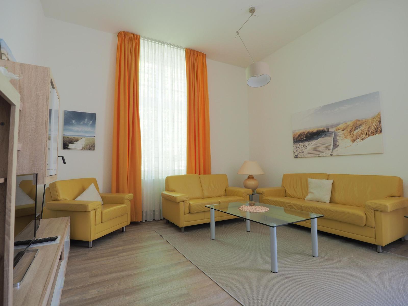 Wohnzimmer mit gelben Sitzmöbeln, Glas-Tisch und orangefarbenen Vorhängen.