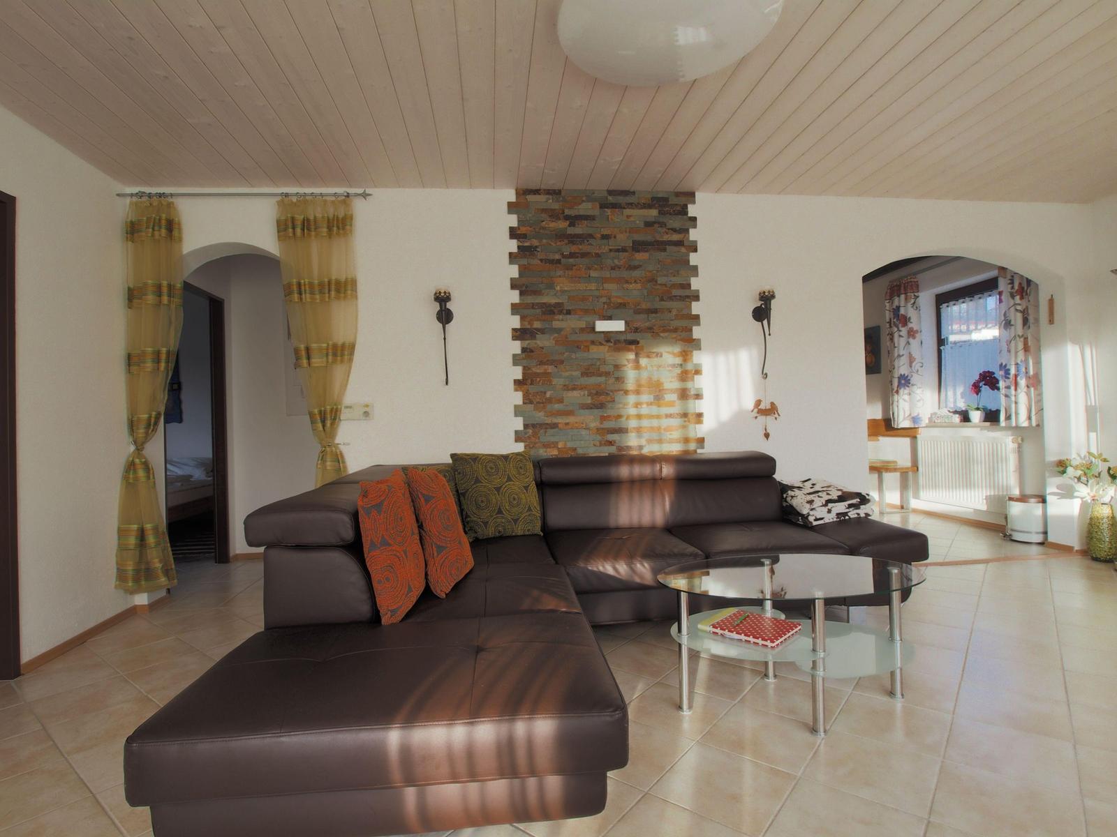 Living room with brown sofa, glass table, and stone wall. Doorways to bedroom and window area.