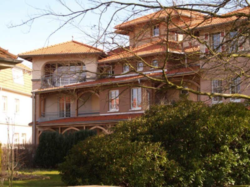 Haus mit rotem Dach und Balkonen, umgeben von Bäumen und Sträuchern.