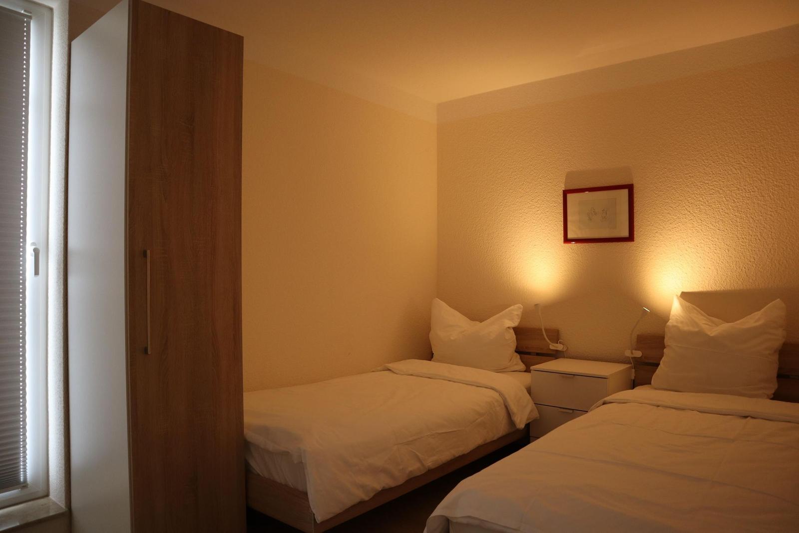 Zwei Betten mit weißen Bettwäsche, Nachttisch und Kleiderschrank neben Fenster.