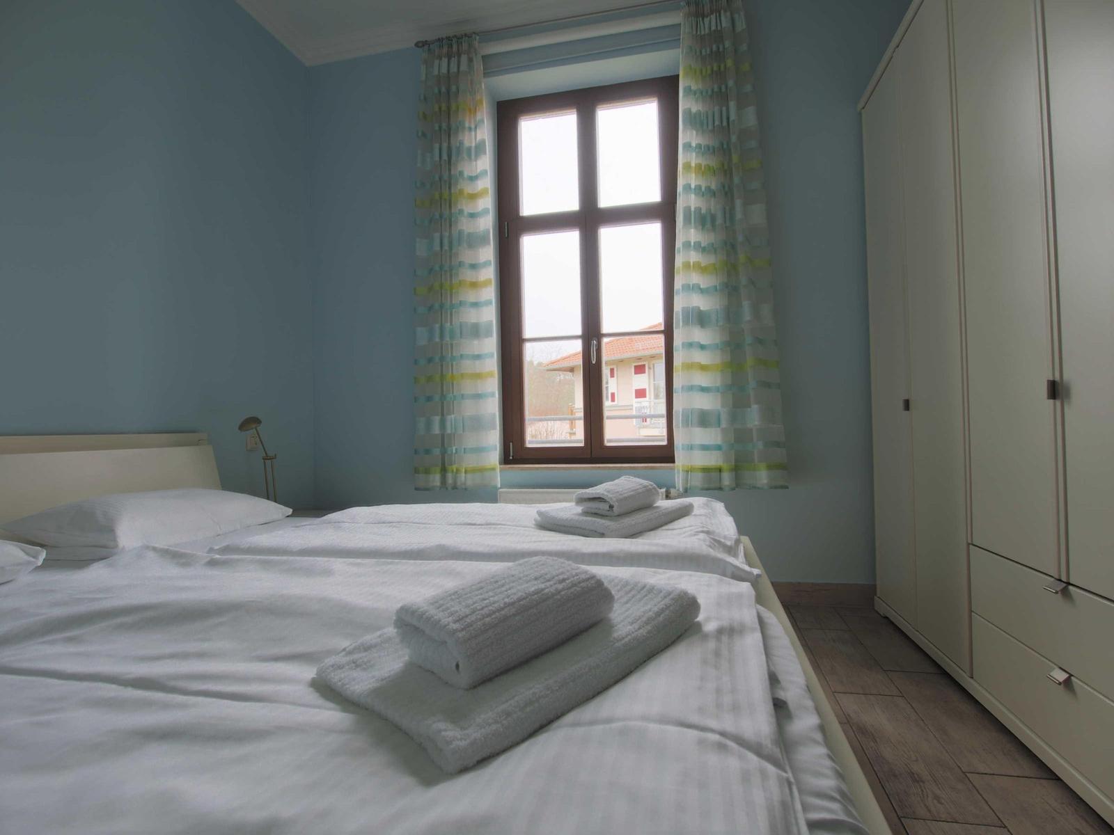 Schlafzimmer mit Bett, Gardinen und Kleiderschrank. Fensterblick auf Gebäude.