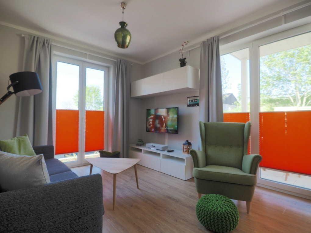 Wohnzimmer mit Sofa, Sessel und Fernseher. Große Fenster mit orangefarbenen Jalousien.