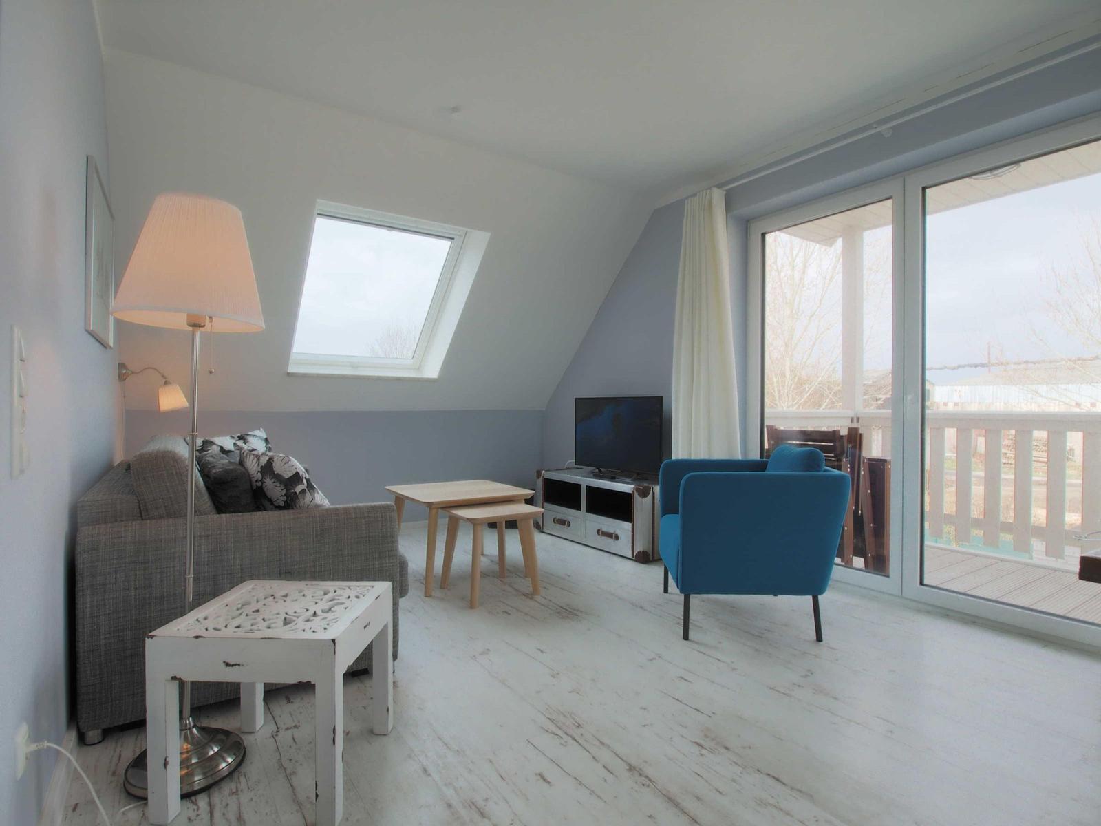 Wohnzimmer mit Sofa, blauem Sessel, TV und Balkonzugang.