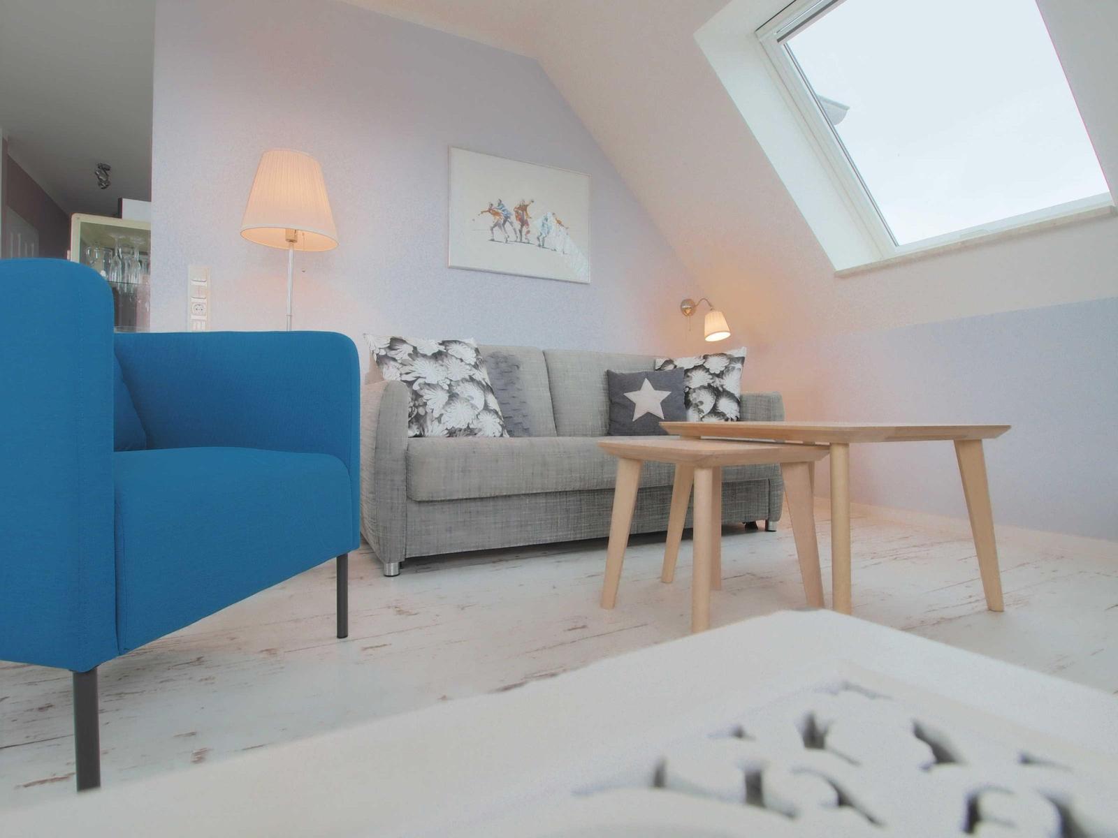 Ein Wohnzimmer mit grauem Sofa, blauem Sessel und Holztisch unter einem Dachfenster.
