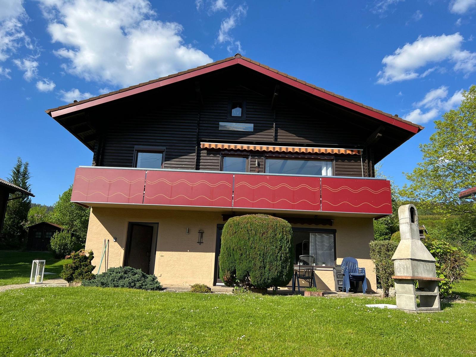 Two-story house with balcony, garden, and grill area under blue sky.