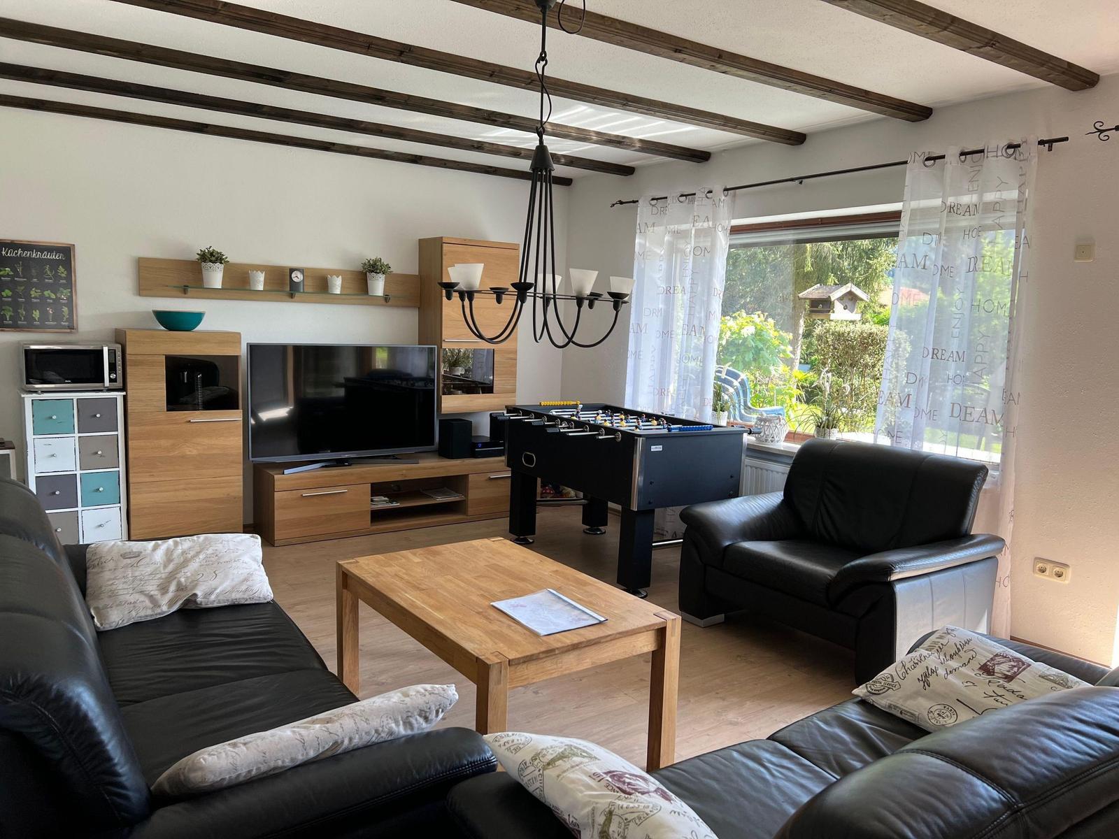 Living room with sofa, table, TV, and foosball table. Wooden beams and window with curtains.