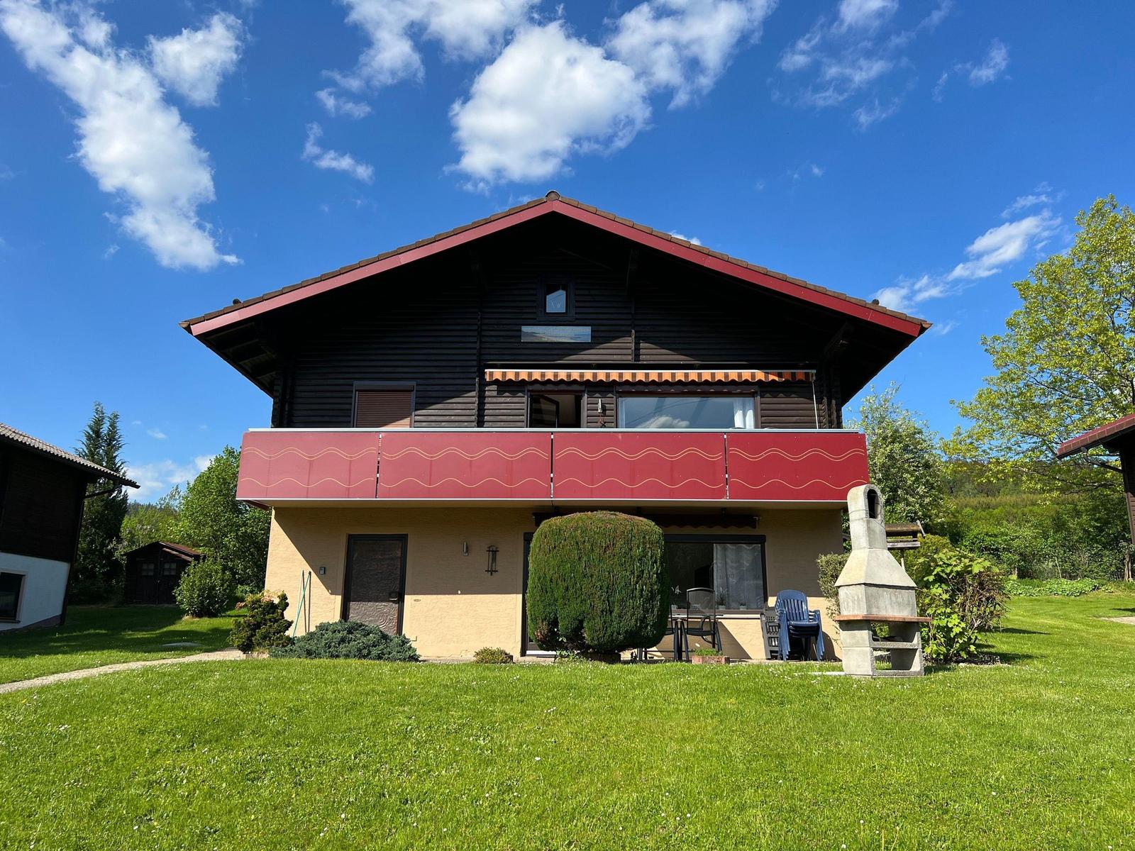 House with balcony, garden, and grill area under blue sky.