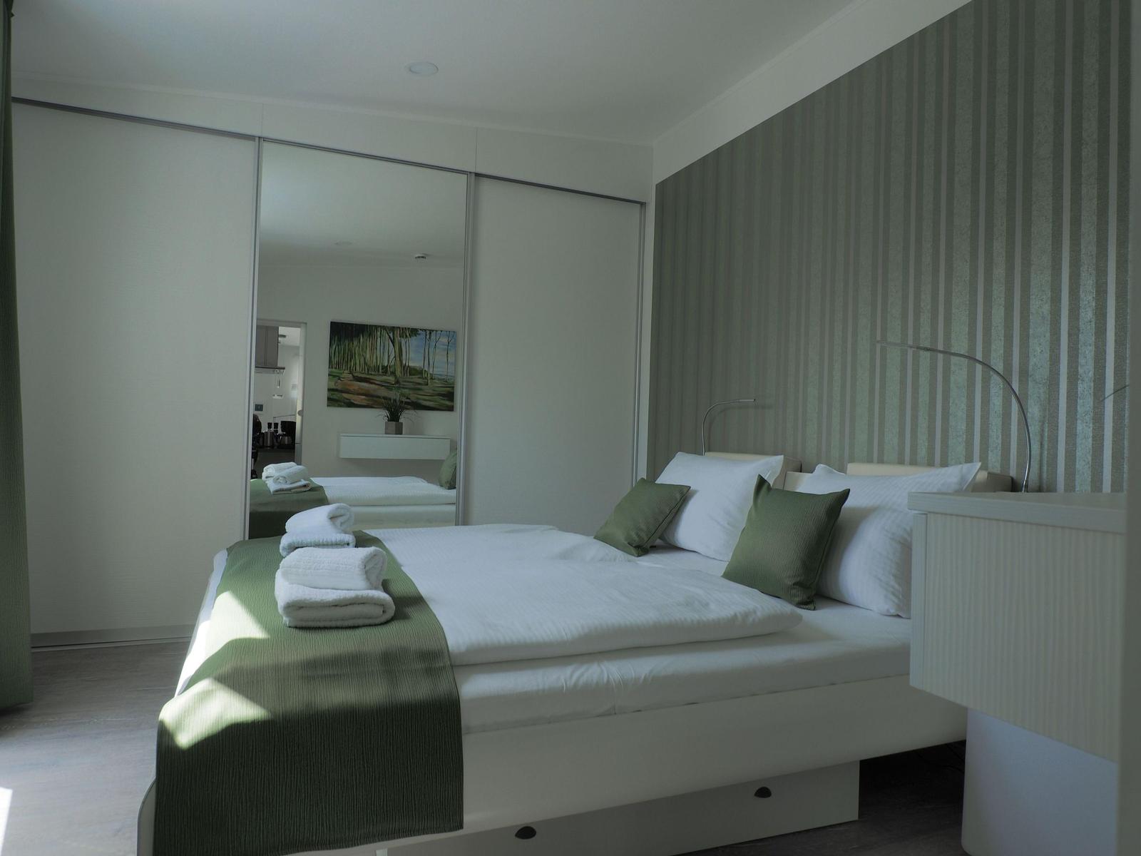 Schlafzimmer mit Bett, grünen Kissen und Spiegelschrank. Wandtapete mit Streifenmuster.