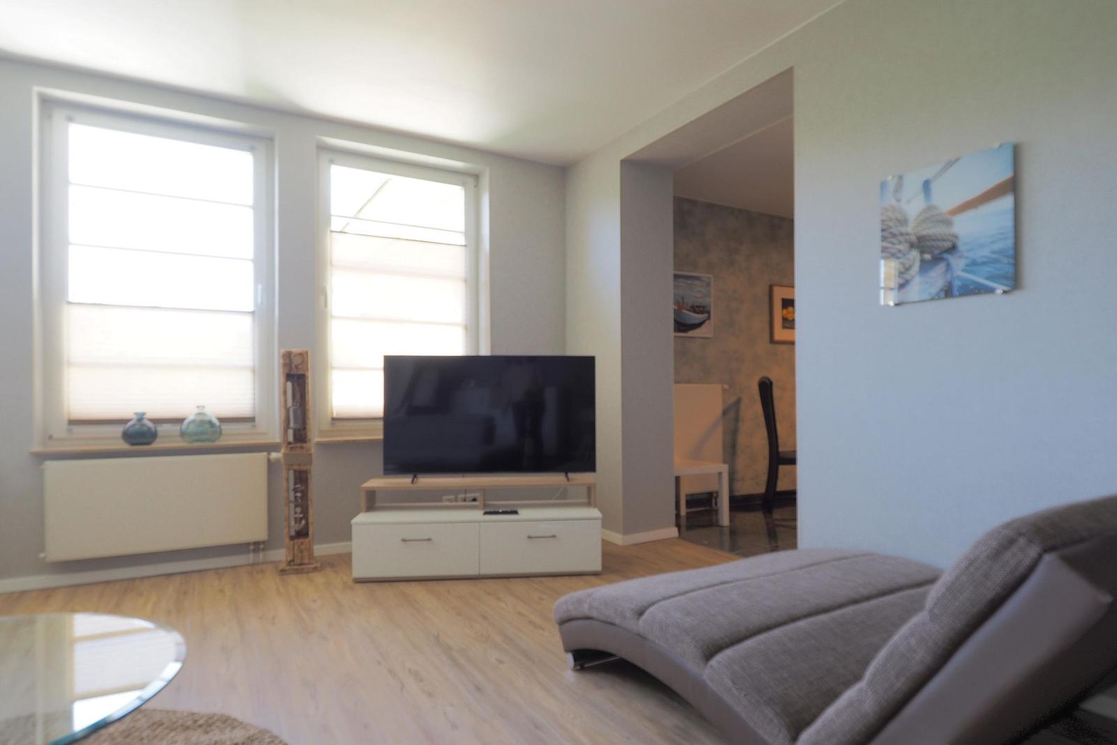 Wohnzimmer mit Sofa, Fernseher und Fenstern. Blick in die Küche.
