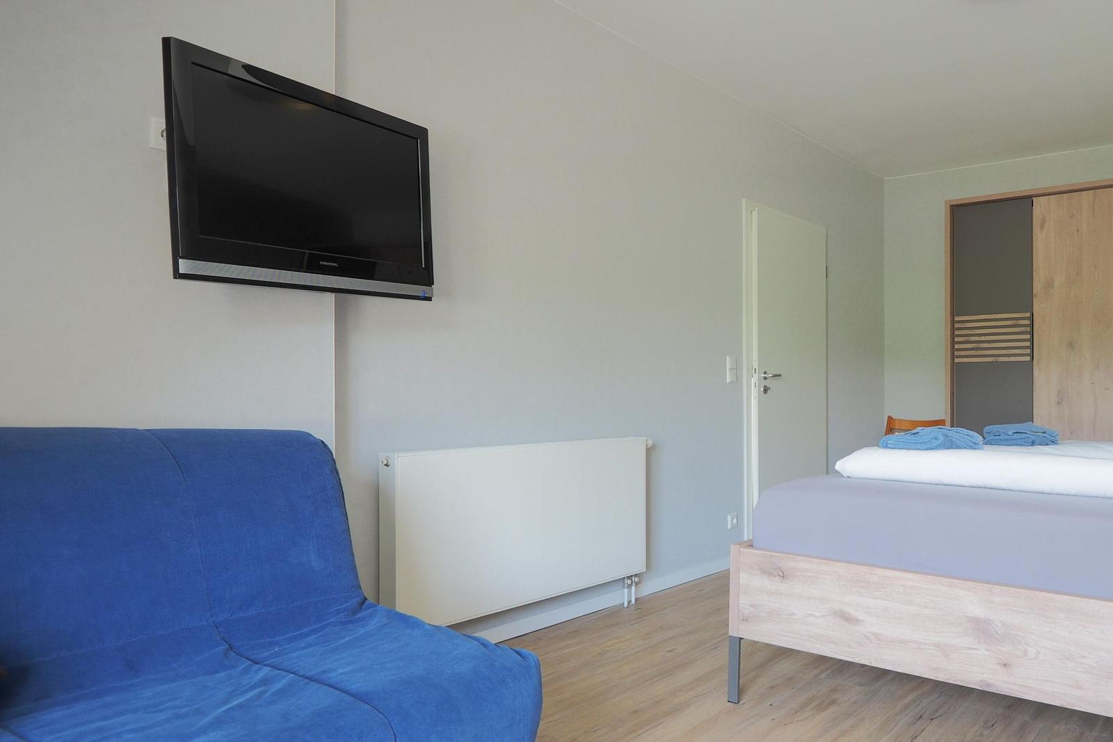 Zimmer mit Bett, Sofa und Wandfernseher. Holzboden und Kleiderschrank sichtbar.