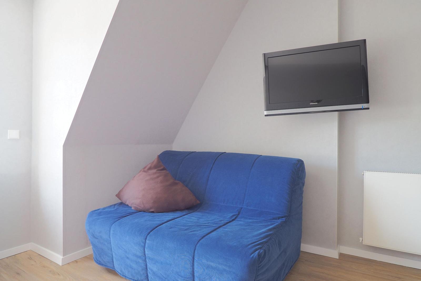 Ein blauer Sessel mit Kissen unter einer schrägen Decke. An der Wand hängt ein Fernseher.