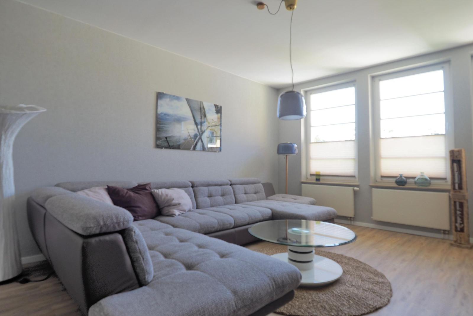 Ein Wohnzimmer mit grauem Sofa, Glas-Tisch und Fenstern mit Jalousien.