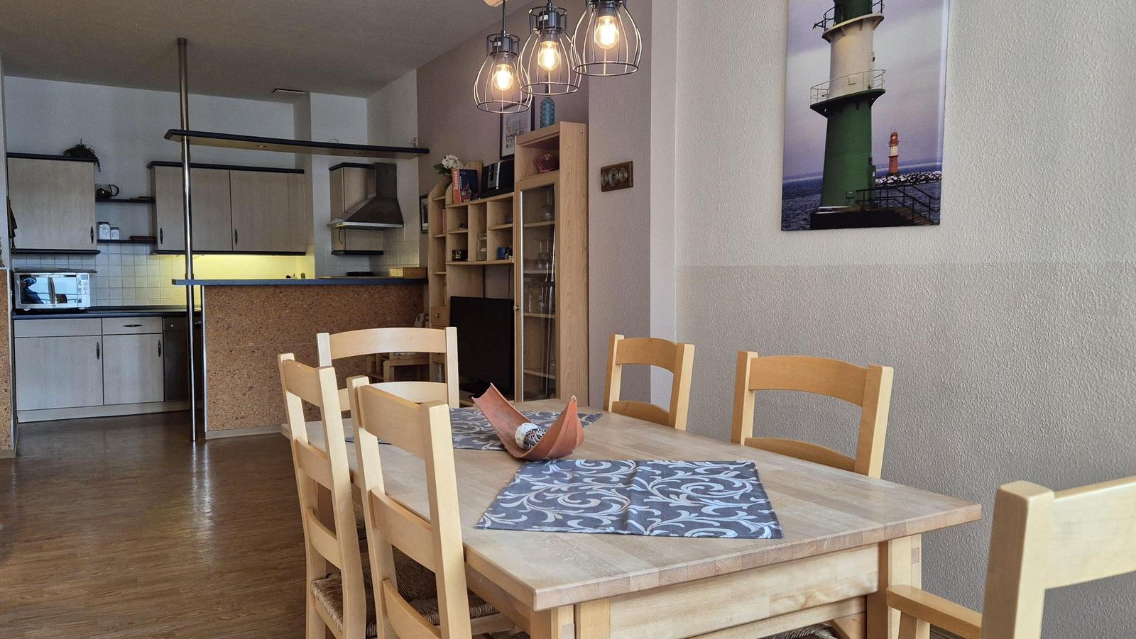 Essbereich mit Holztisch und Stühlen, Küchenbereich im Hintergrund, Wandbild eines Leuchtturms.