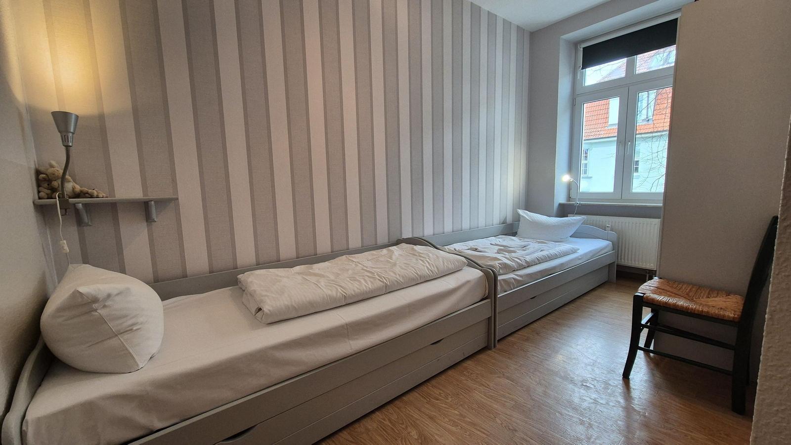 Zimmer mit zwei Einzelbetten, gestreifter Wand und Holzboden. Fenster mit Blick auf Gebäude.