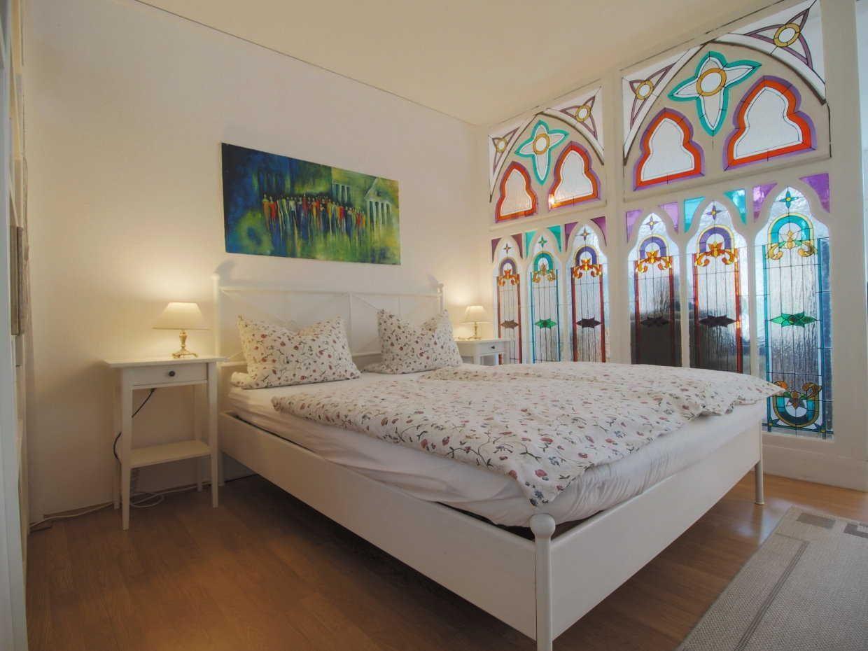 Ein Schlafzimmer mit weißem Bett, Nachttischen und bunten Glasfenstern.
