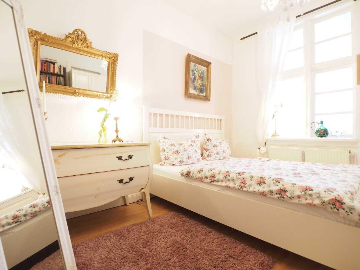 Schlafzimmer mit Bett, Schrank und Spiegel an der Wand. Fenster mit Vorhängen.