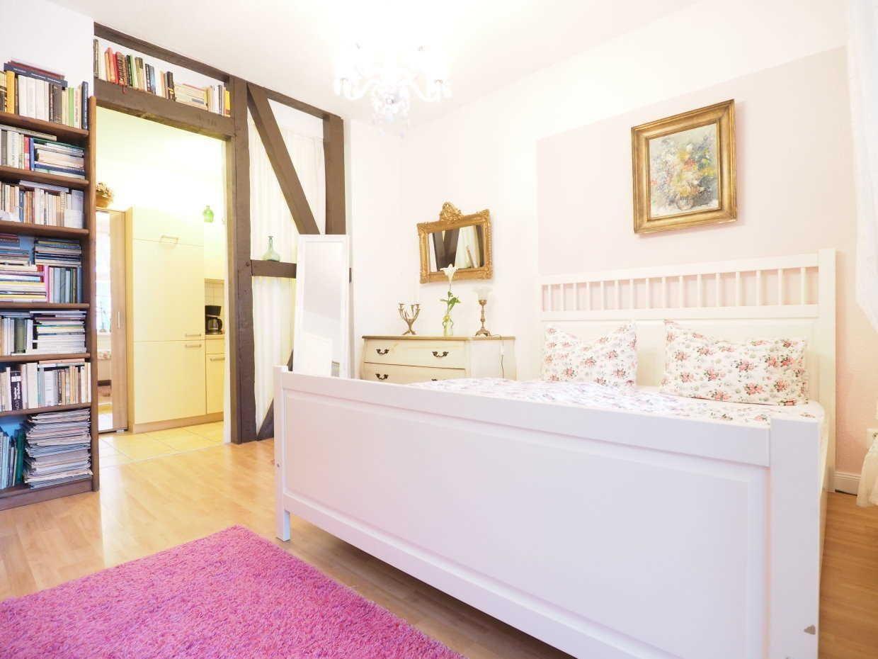 Schlafzimmer mit weißem Bett, rosa Teppich und Bücherregal. Durchgang zu Küche mit Holzrahmen.