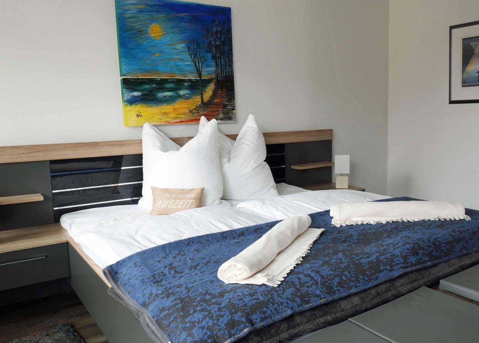Doppelbett mit blauer Decke, weißen Kissen und Wandbild