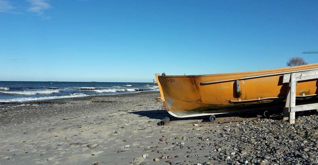 Ein orangefarbener Bootsrumpf steht auf einem steinigen Strand vor dem Meer.