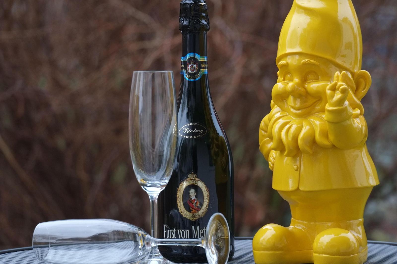 Auf einem Tisch steht eine Flasche Sekt, ein Glas und eine gelbe Gnome-Figur.
