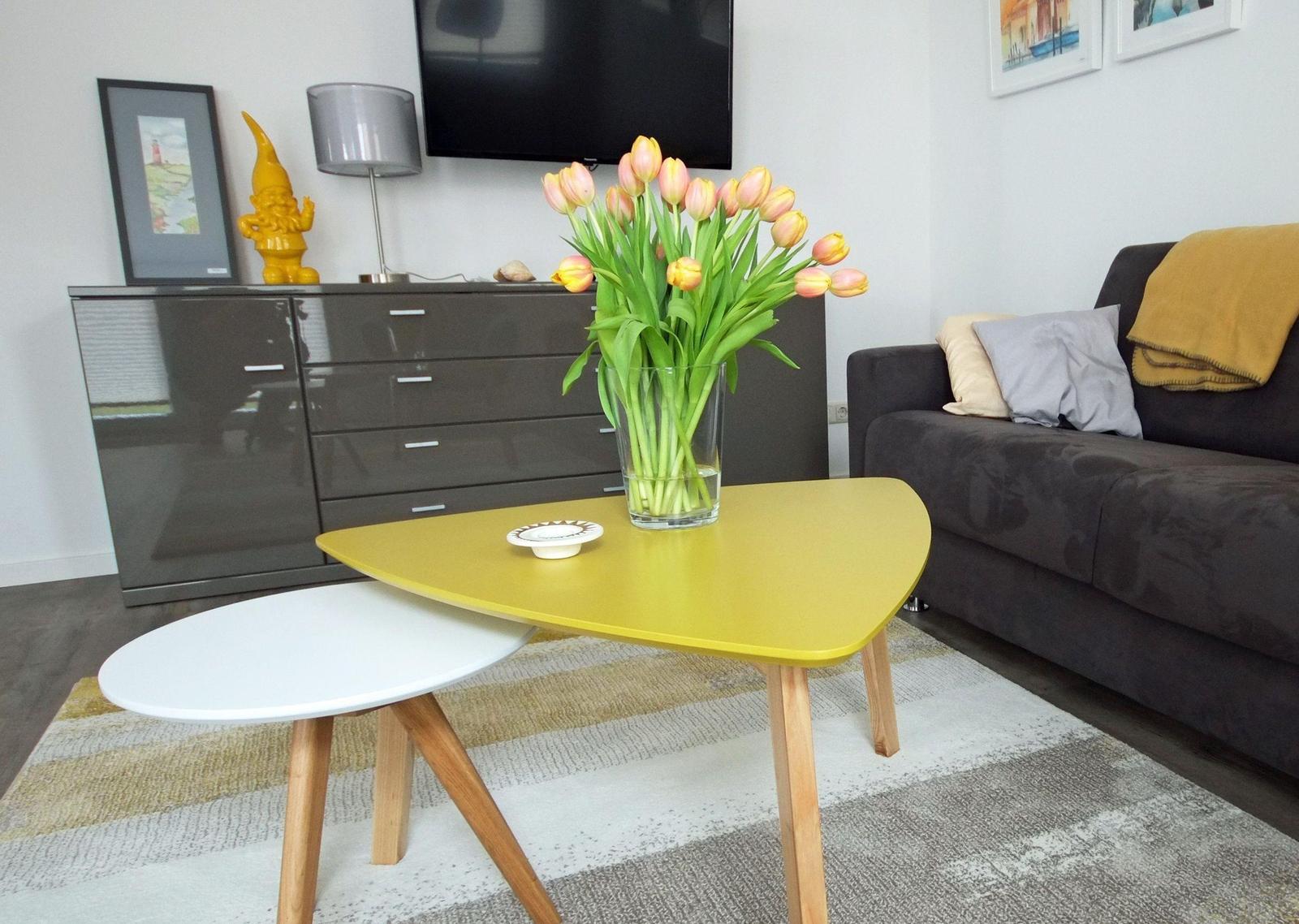 Ein Wohnzimmer mit Sofa, Couchtisch und Blumen. Hintergrund: Schrank und Fernseher.