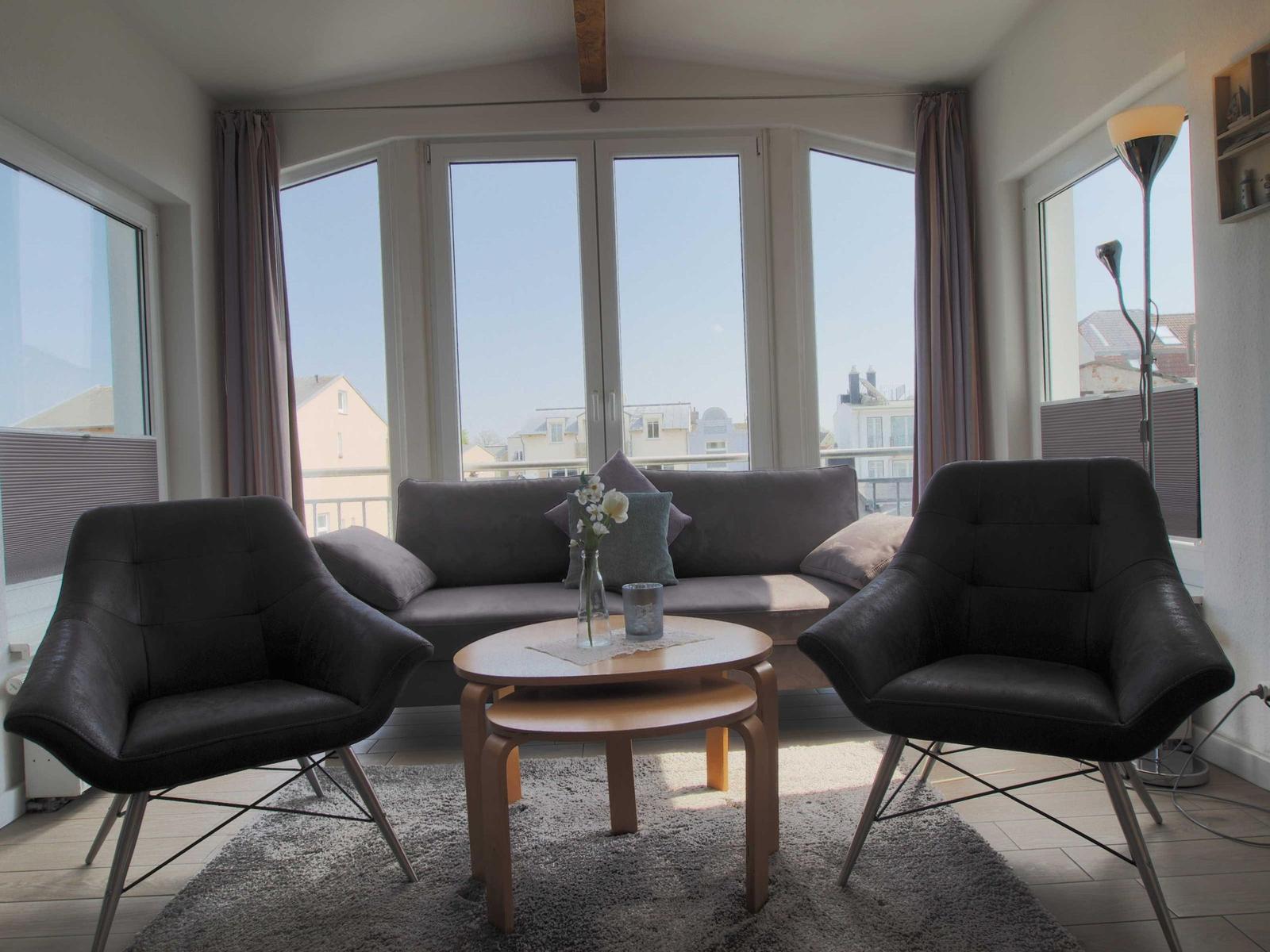 Ein Wohnzimmer mit Sofa, zwei Sesseln und einem runden Tisch. Große Fenster bieten Blick auf die Stadt.