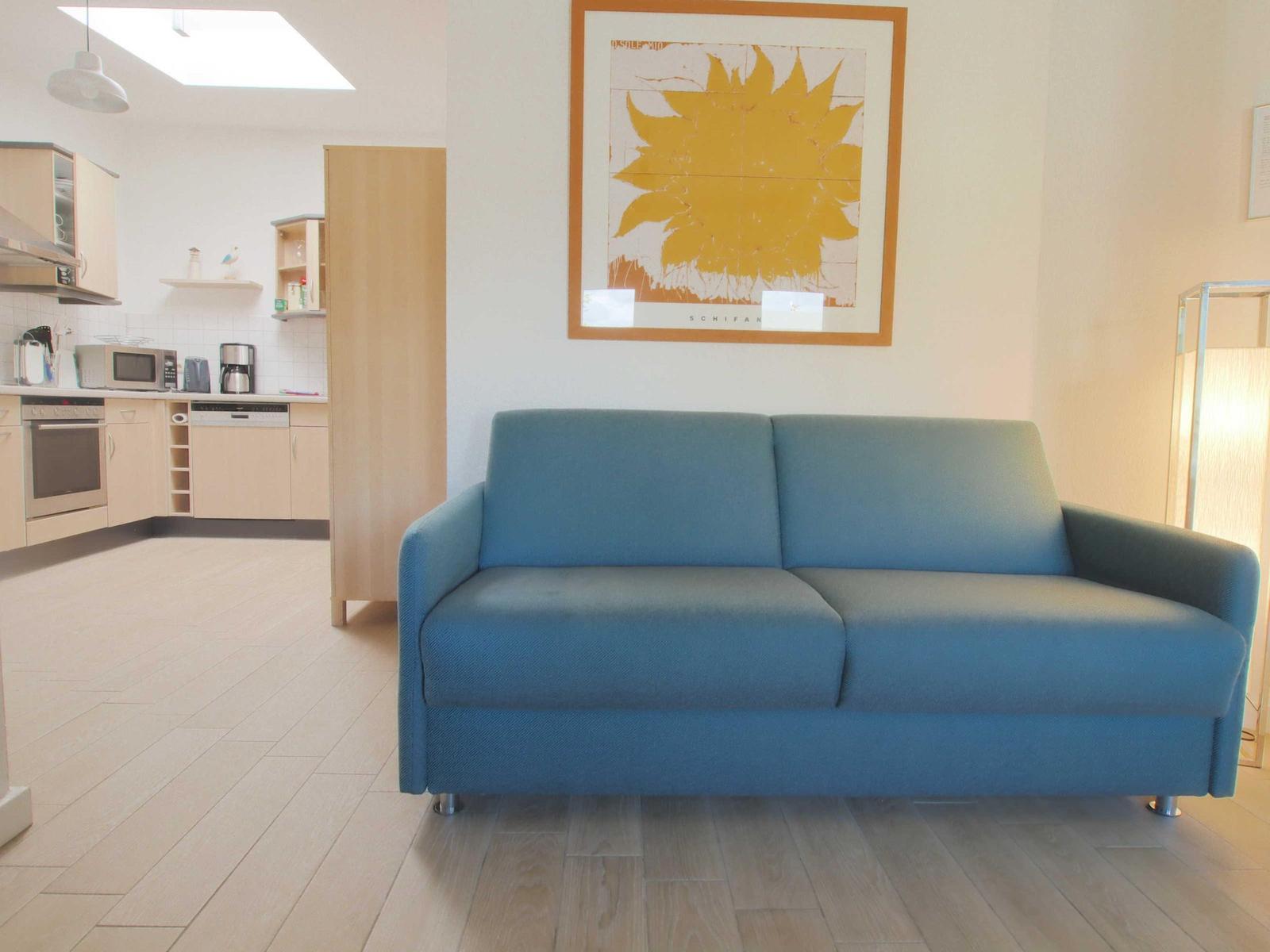 Blauer Sofa im Wohnbereich mit Küchenbereich im Hintergrund und Wandbild.