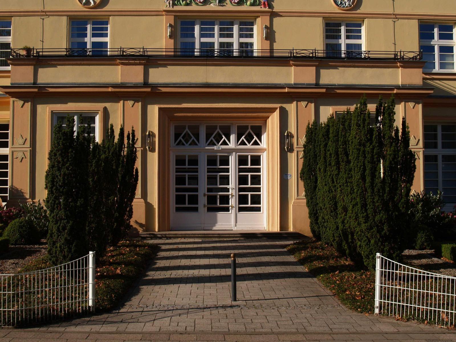 Eingangsbereich mit weißen Türen und Zierhecken vor einem hellen Gebäude.