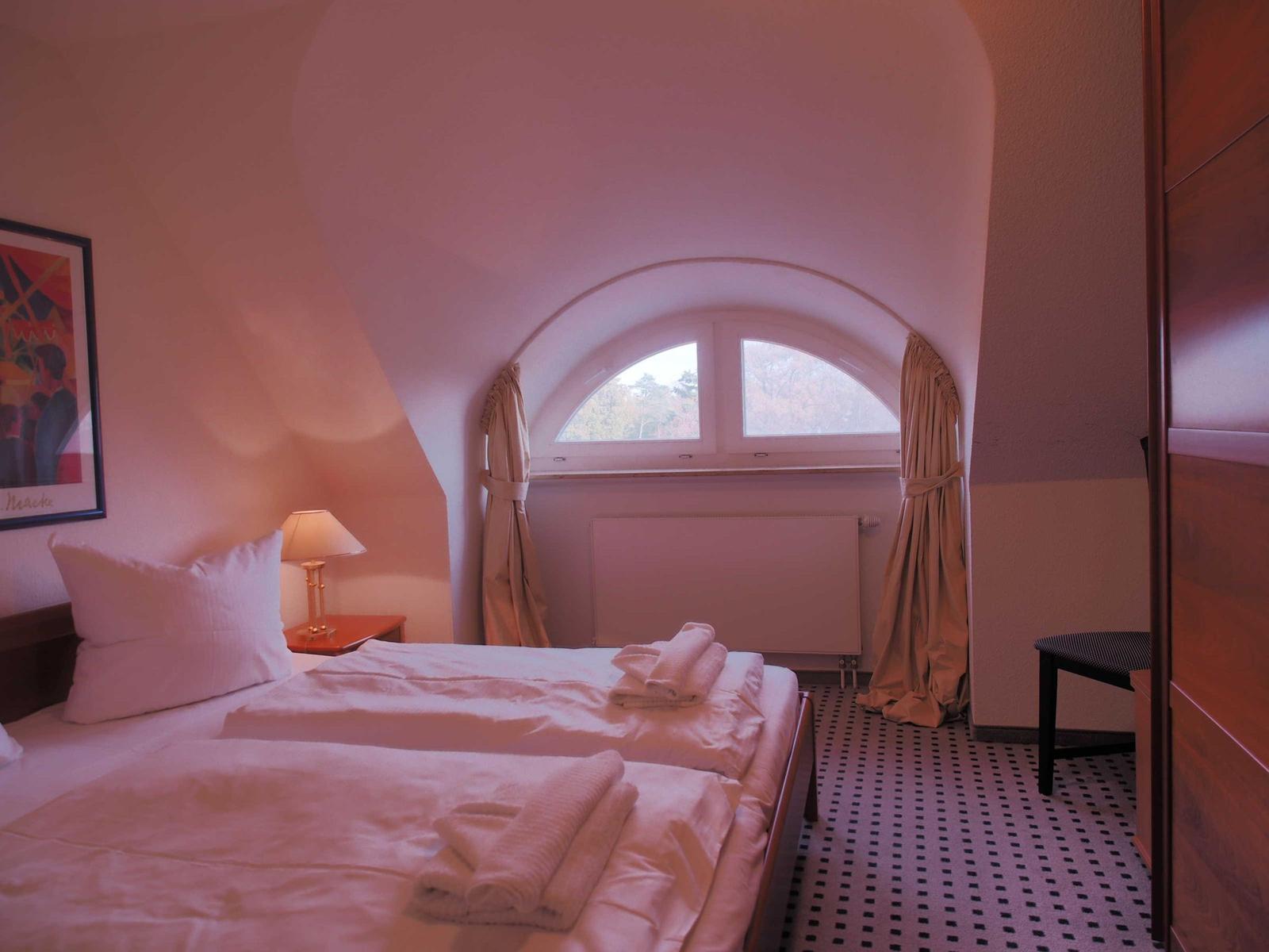Schlafzimmer mit Doppelbett, Bogenfenster und Holzschrank. Bodenfliesen und Wandbild sichtbar.