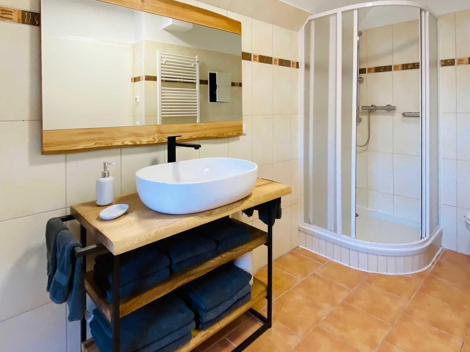 Badezimmer mit Dusche, Waschtisch und Spiegel. Holzregal mit Handtüchern.